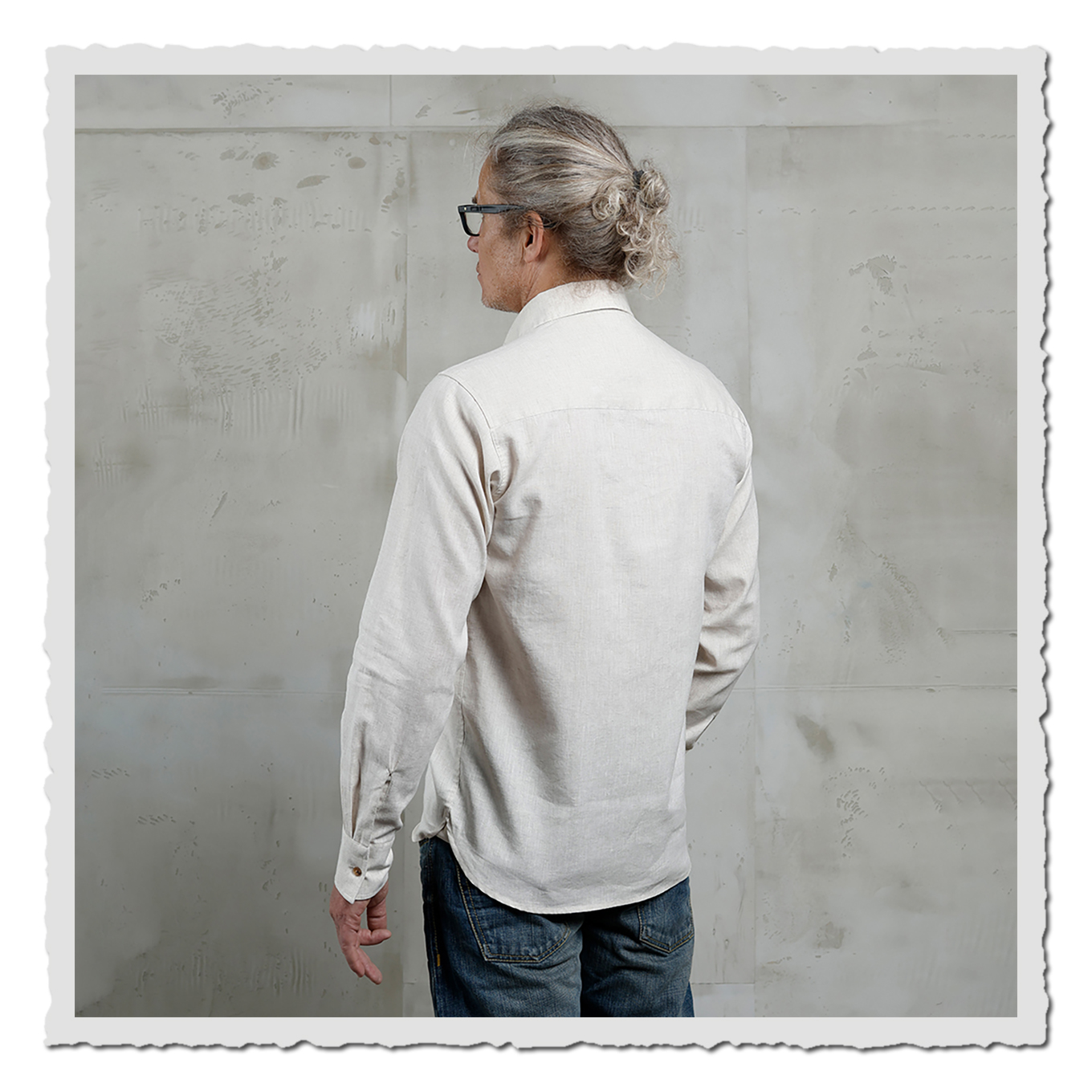 Rückenansicht Model im naturweißen Langarmhemd, seitlich gedreht, grauer Betonhintergrund.